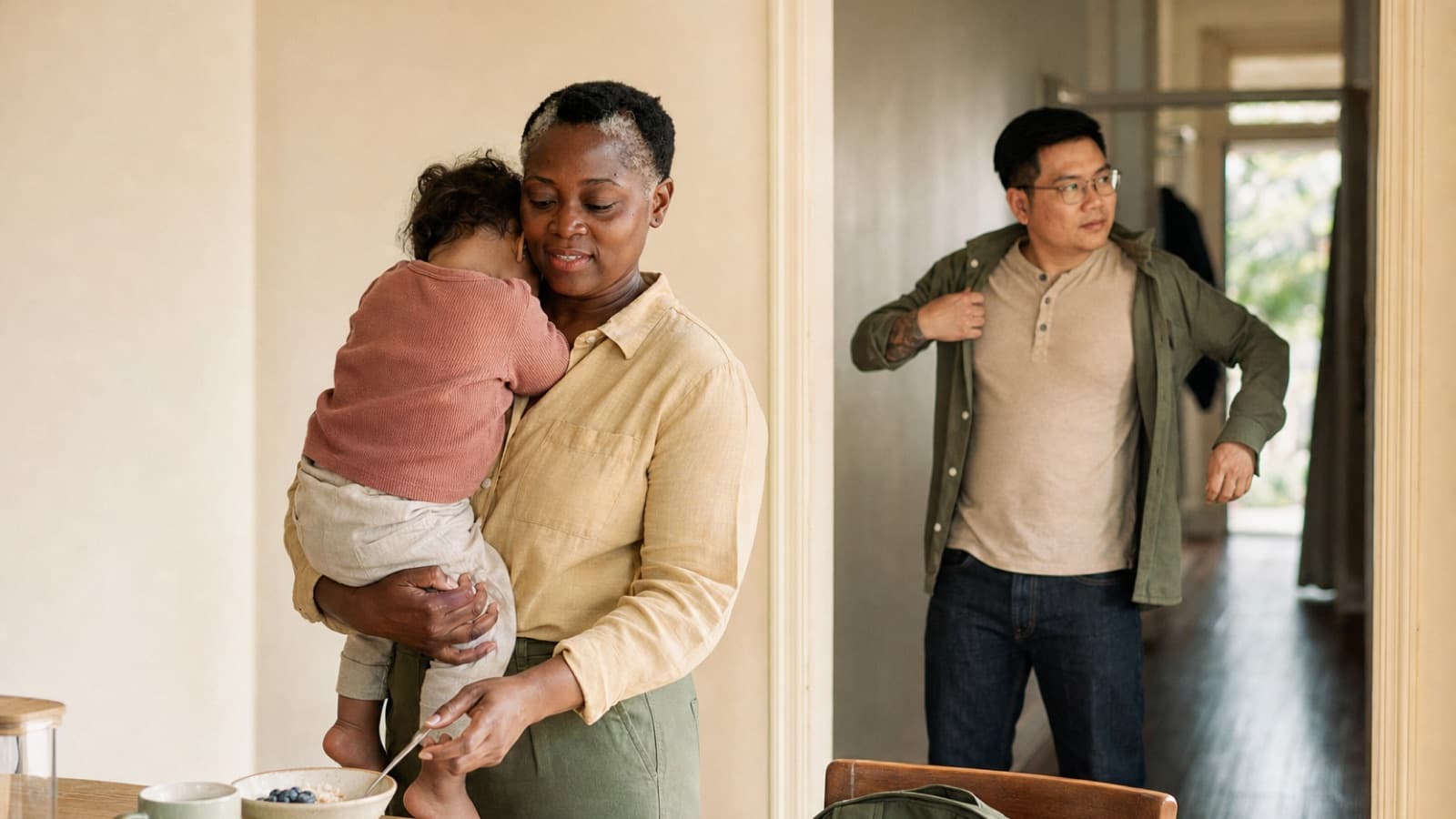 A nanny holding a toddler in a kitchen as a parent leaves for the day